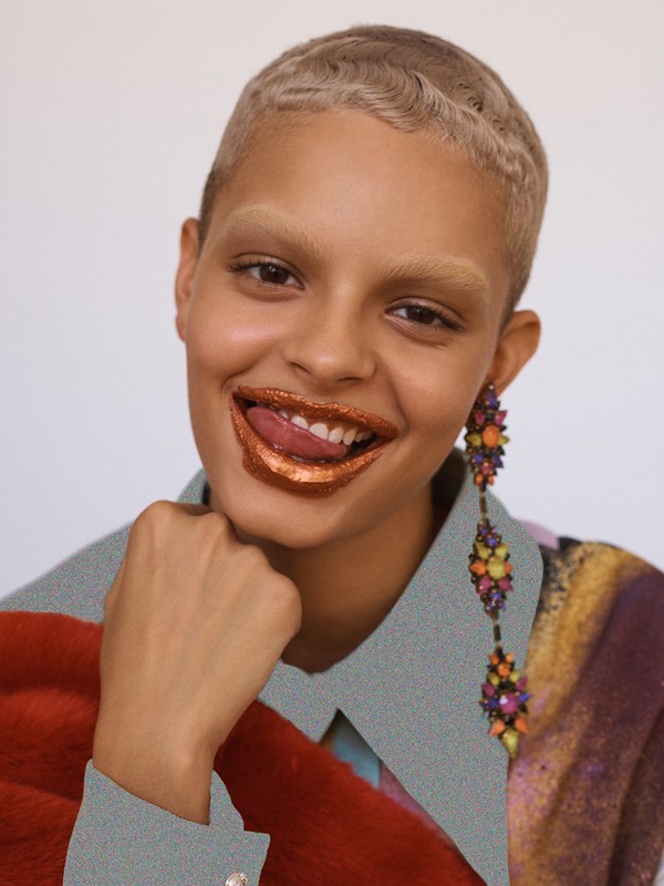 Close-up of a young adult woman with short hair and glittering gold lipstick, posing with a playful expression in a colorful outfit.