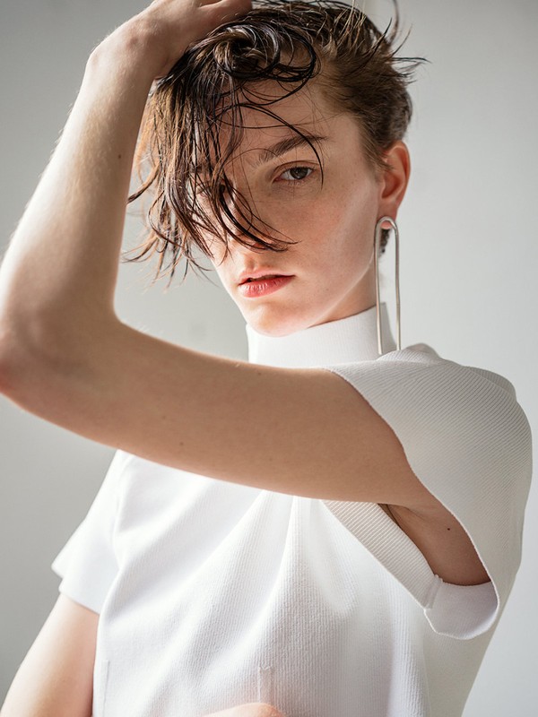 Portrait of a young adult female model with wet short hair and large silver earrings in a white top, posing against a white wall.