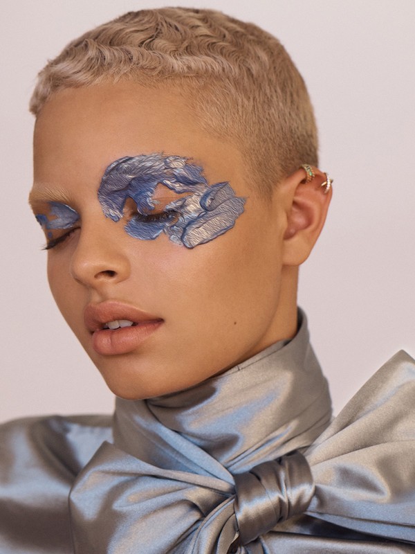 Close-up of a young adult woman with fair skin and short blonde hair, wearing blue and silver eye makeup and a satin ribbon-bow top.