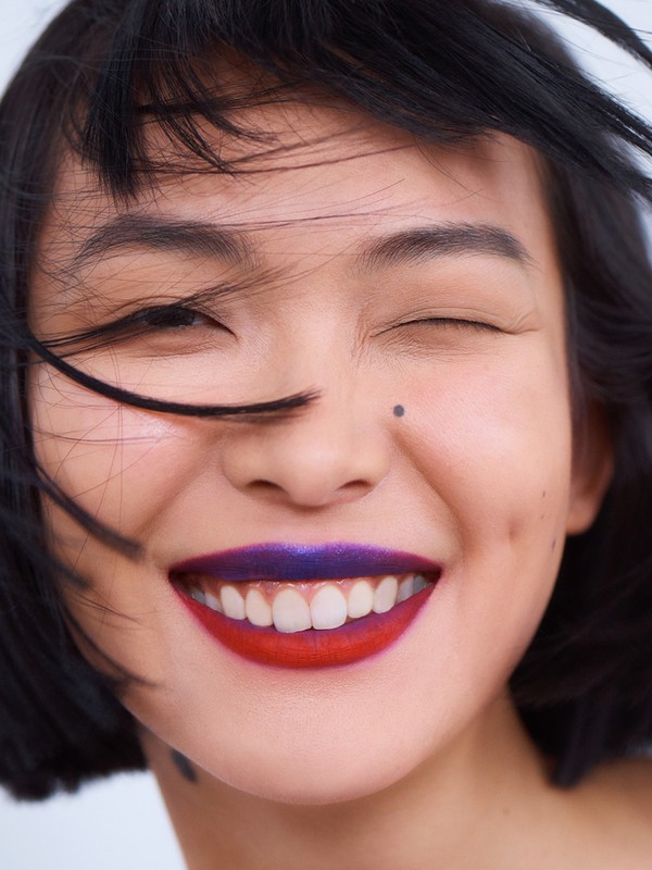 Close-up of a smiling Asian woman with short black hair and two-tone lipstick, one eye closed, against a white background.