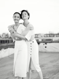 Two smiling women in white outfits embracing outdoors with a bright sky in the background