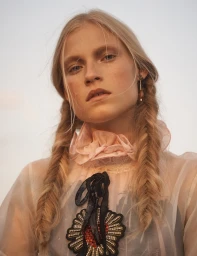 Close-up portrait of a blonde woman with braided pigtails wearing a sheer ruffled blouse with beaded floral pendant necklace.