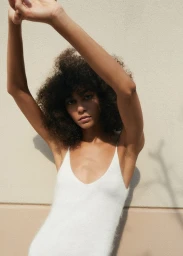Model with dark curly hair wearing a white textured tank top, posing with arms raised against a plain wall.