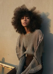 A woman with large voluminous curly hair wearing a metallic ribbed sweater leaning against a concrete wall outdoors.