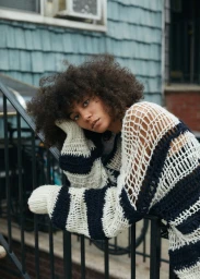 A woman with voluminous dark curly hair leans over a black metal staircase, wearing an oversized striped knit sweater.