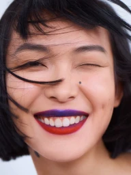 Close-up portrait of a smiling woman with dark hair blowing across her face, vivid purple and red lipstick, and a distinct beauty mark on her cheek.