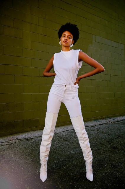 Stylish young Black woman posing confidently against a textured green brick wall, wearing a white top, white jeans, and high white boots.
