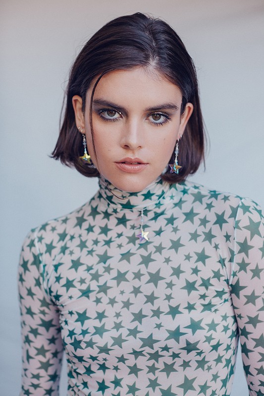 A young woman with bobbed hair poses indoors wearing a sheer top with green star patterns, statement earrings, and smokey eye makeup.