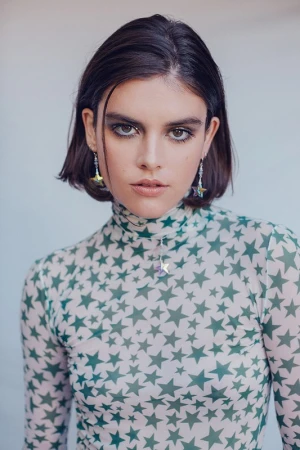 A young woman with bobbed hair poses indoors wearing a sheer top with green star patterns, statement earrings, and smokey eye makeup.