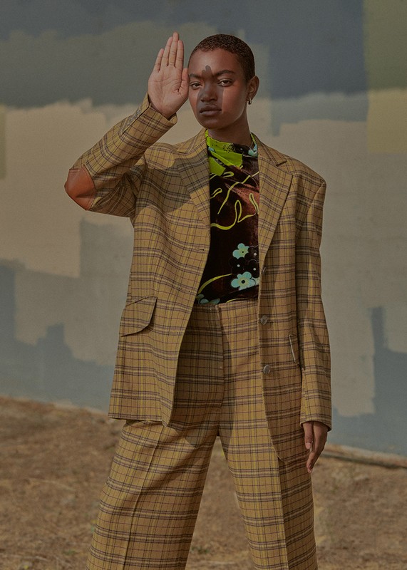 A young adult woman poses outdoors in a tan plaid suit against a painterly backdrop of muted blues and tans in a fashion editorial.