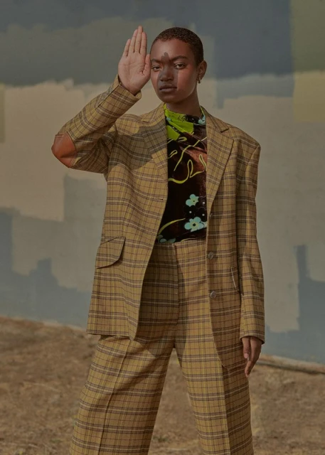 A young adult woman poses outdoors in a tan plaid suit against a painterly backdrop of muted blues and tans in a fashion editorial.