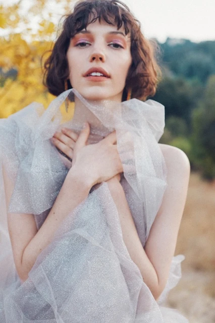 Adult female with curly brown hair and pink eyeshadow poses in sheer grey dress in an outdoor setting with trees and yellow leaves.