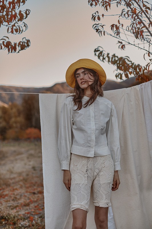 A woman in a straw hat and white blouse stands outdoors, with a background of autumn leaves and a draped white cloth. Fashion editorial.