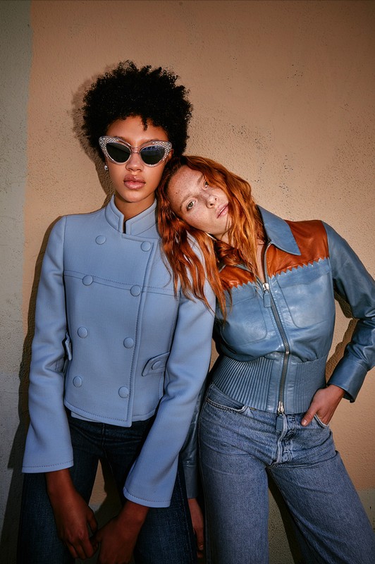 Two women pose in fitted jackets and jeans against a neutral wall. One has dark curly hair and jeweled sunglasses, and the other has red hair.