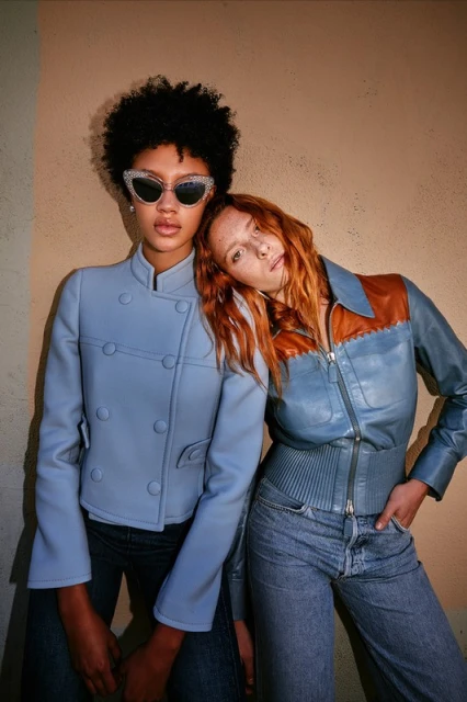 Two women pose in fitted jackets and jeans against a neutral wall. One has dark curly hair and jeweled sunglasses, and the other has red hair.