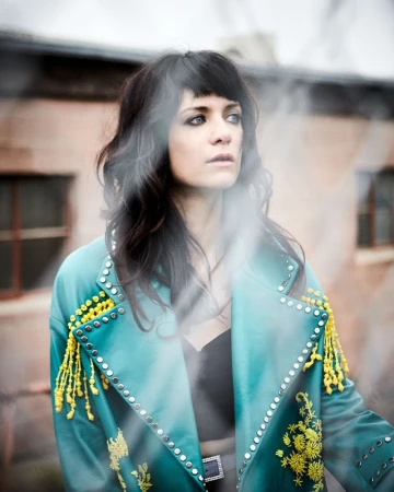 Brunette woman with bangs in a teal coat with yellow tassels stands outdoors in front of a brick building, gazing off to the side.
