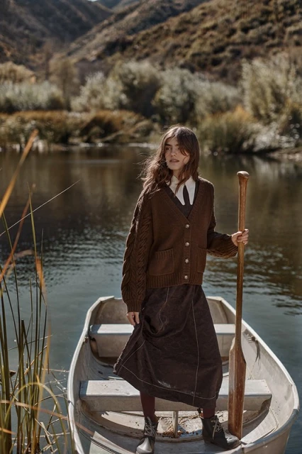 Woman stands in a boat with a paddle on a lake in front of mountains. She is wearing a cardigan, dress, and boots.