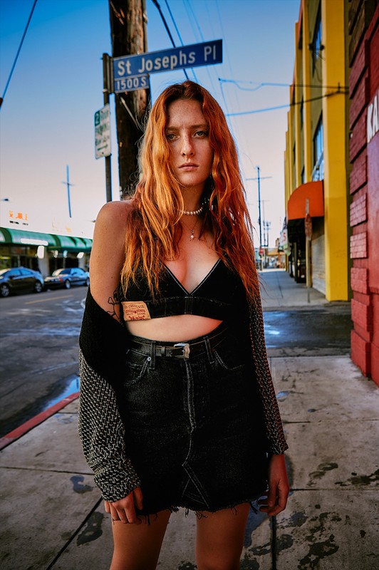 A young adult woman with red hair poses on an urban street wearing a black denim outfit with a grey sweater and buildings in the background.