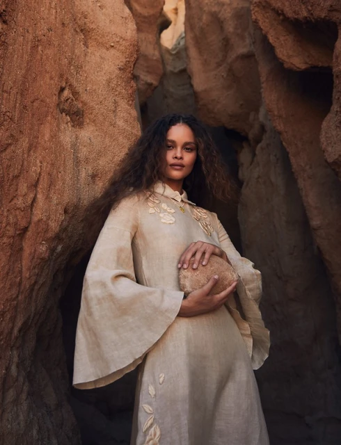 A woman with curly hair poses with a small boulder in a flowing beige dress, set against a backdrop of light brown rock formations.
