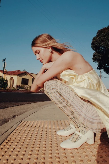 A young woman with blond hair wearing a yellow dress and patterned tights sits on a sidewalk next to a road on a sunny day.