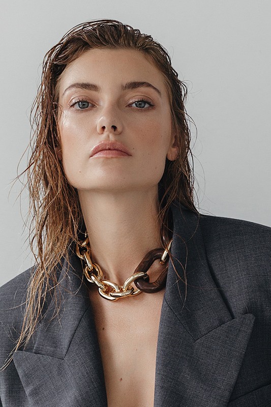 Close-up portrait of a woman with wet hair wearing a gold and brown chunky chain necklace and an oversized grey blazer against a grey background.