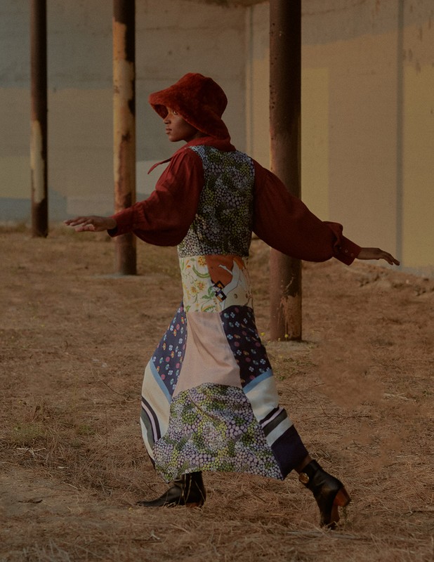 Woman poses outdoors in a patchwork skirt, rust-colored top and hood, and black boots. The background features weathered architecture.