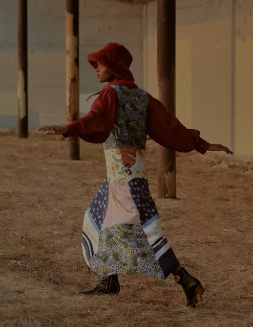 Woman poses outdoors in a patchwork skirt, rust-colored top and hood, and black boots. The background features weathered architecture.