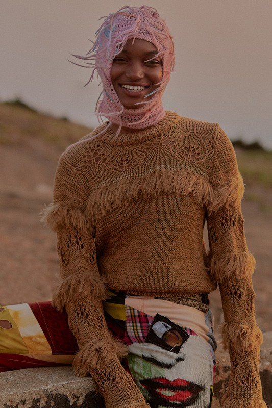 A smiling Black woman in a knitted cap and sweater sits outdoors. Earthy tones and soft lighting create a warm, relaxed fashion editorial.