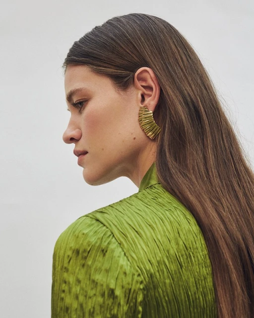 Profile of a woman with long brown hair wearing a green textured top and statement gold earring against a neutral background.