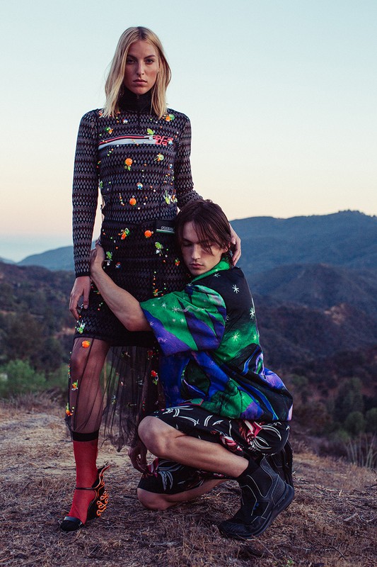 Two fashion models pose outdoors on a hilltop wearing patterned tops, skirts, and boots against a backdrop of mountains and sky.
