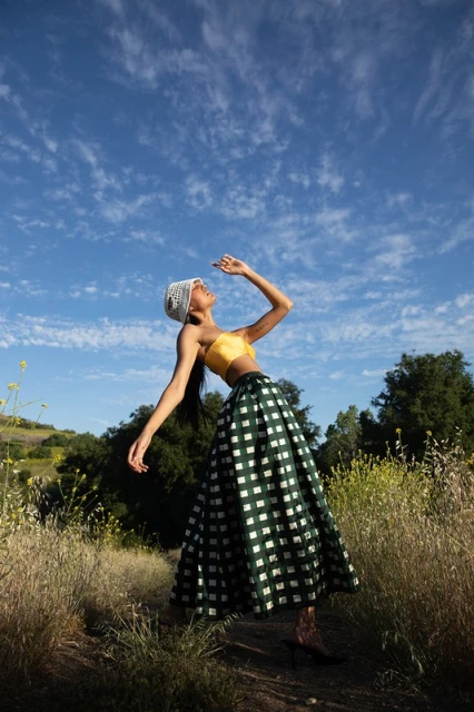 Woman in yellow top and green plaid skirt poses in sunlit field under blue sky with wispy clouds. Summer fashion, outdoor photoshoot.
