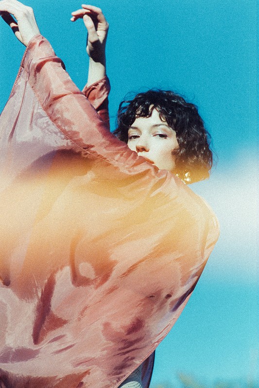 Adult woman in a flowing peach-colored garment poses against a clear blue sky, with sunlit curly hair and an artistic, dreamy aesthetic.