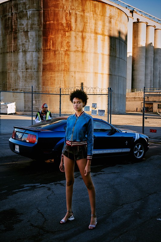 Stylish woman poses in jean jacket and shorts with a blue Mustang car and industrial silos in the background on a sunny day.