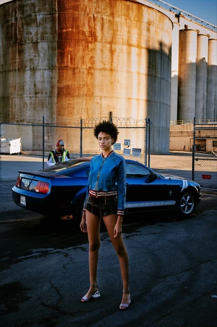 Stylish woman poses in jean jacket and shorts with a blue Mustang car and industrial silos in the background on a sunny day.