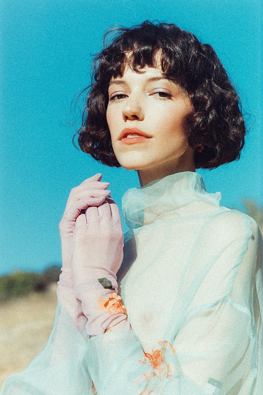 Woman in sheer floral top with pink gloves poses outdoors against a blue sky, showcasing a soft, ethereal aesthetic and natural light.
