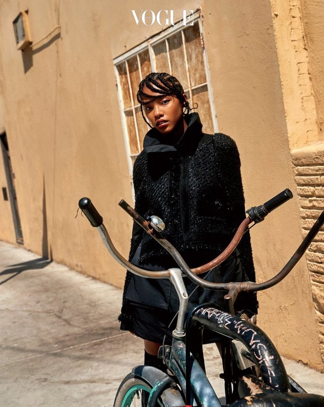 Fashionable woman in a textured black jacket poses next to a vintage bicycle in an urban setting with a Vogue logo in the top corner.