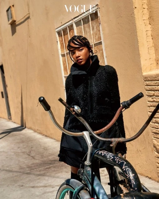 Fashionable woman in a textured black jacket poses next to a vintage bicycle in an urban setting with a Vogue logo in the top corner.