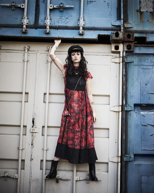 A young woman poses between shipping containers. She wears a dark hat, a patterned dress, and boots, with a small purse over her shoulder.
