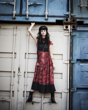 A young woman poses between shipping containers. She wears a dark hat, a patterned dress, and boots, with a small purse over her shoulder.