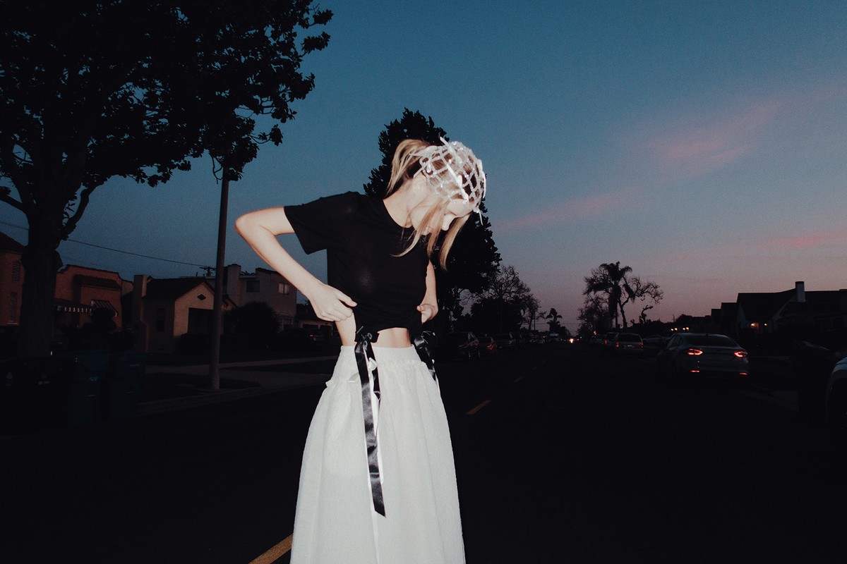 A young adult female wearing a black T-shirt and a long white skirt stands on a dark street near suburban houses at dusk.