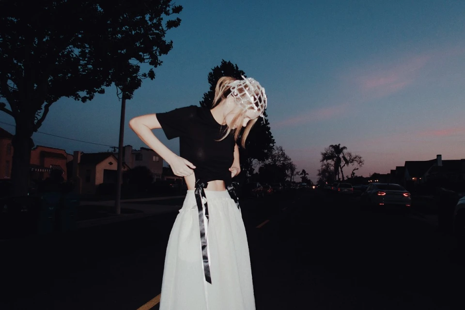 A young adult female wearing a black T-shirt and a long white skirt stands on a dark street near suburban houses at dusk.
