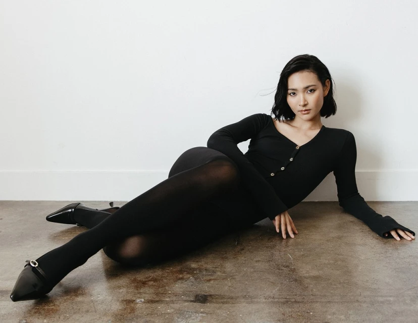 Woman in a black bodysuit and tights reclining on a neutral surface in a studio, showcasing a sleek and fashionable style.