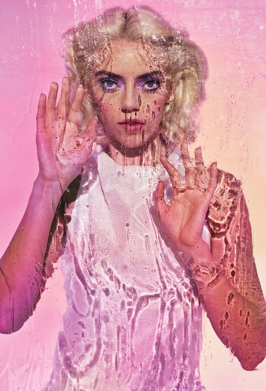 Striking portrait of a young woman with wet hair and water droplets on glass, with cool-toned makeup and a pink and orange background.