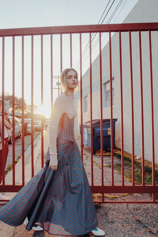 Blonde woman in a long, plaid dress walks by a red metal fence, with a white building and cars in the background.
