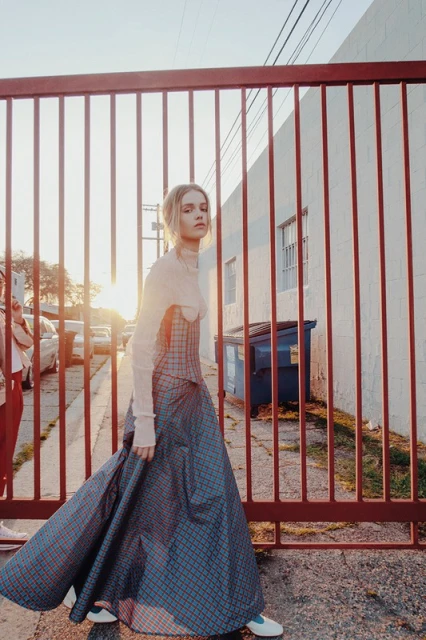 Blonde woman in a long, plaid dress walks by a red metal fence, with a white building and cars in the background.