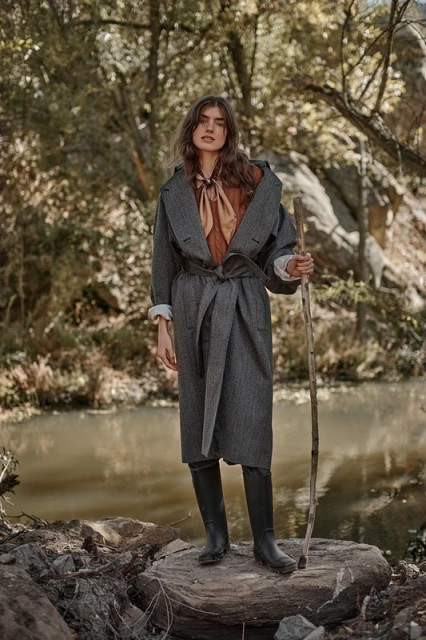 A woman in a grey coat standing on rocks near a river, holding a walking stick, with a forest in the background and diffused lighting.