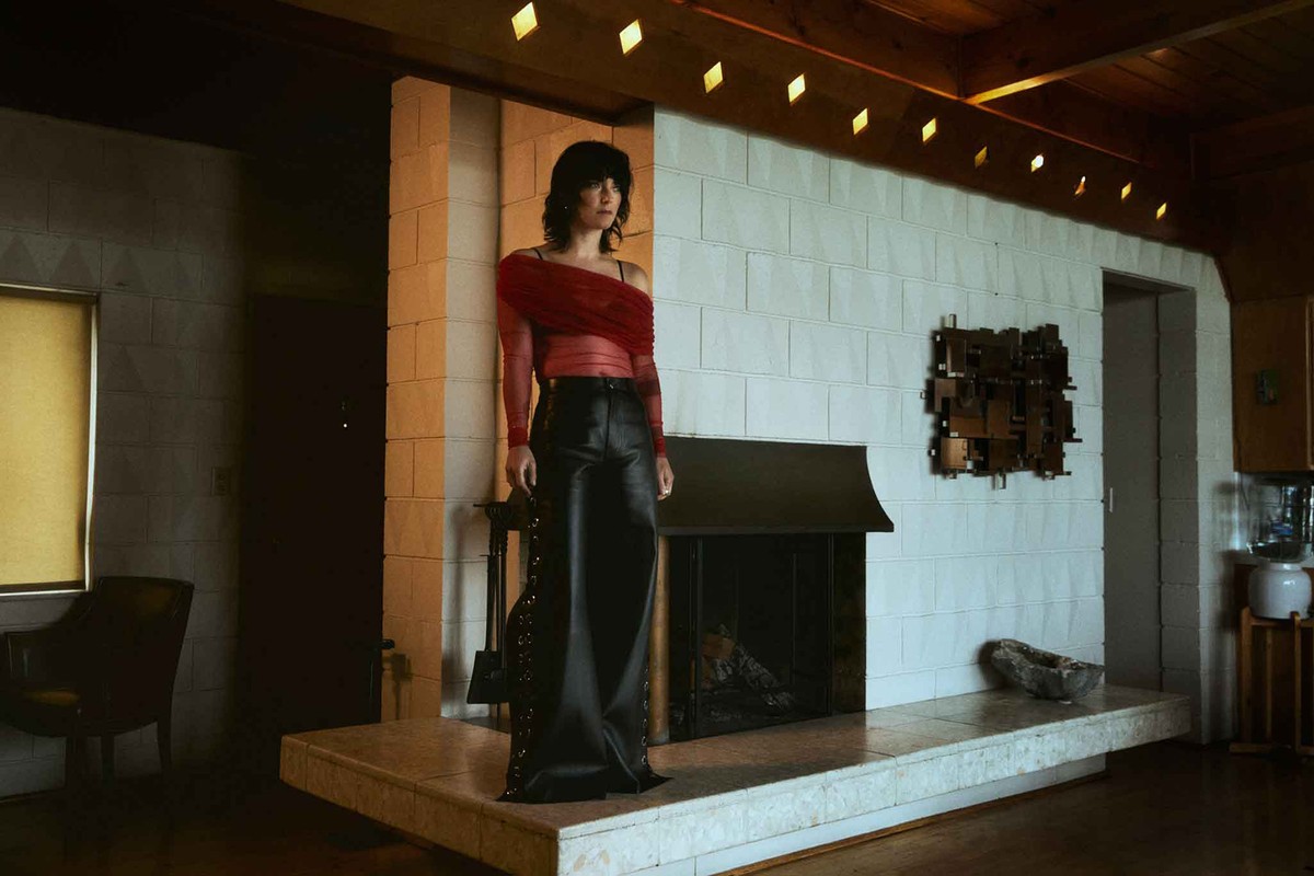 Adult woman posing in a sheer red blouse and black leather pants indoors, standing on a marble platform in front of a fireplace.