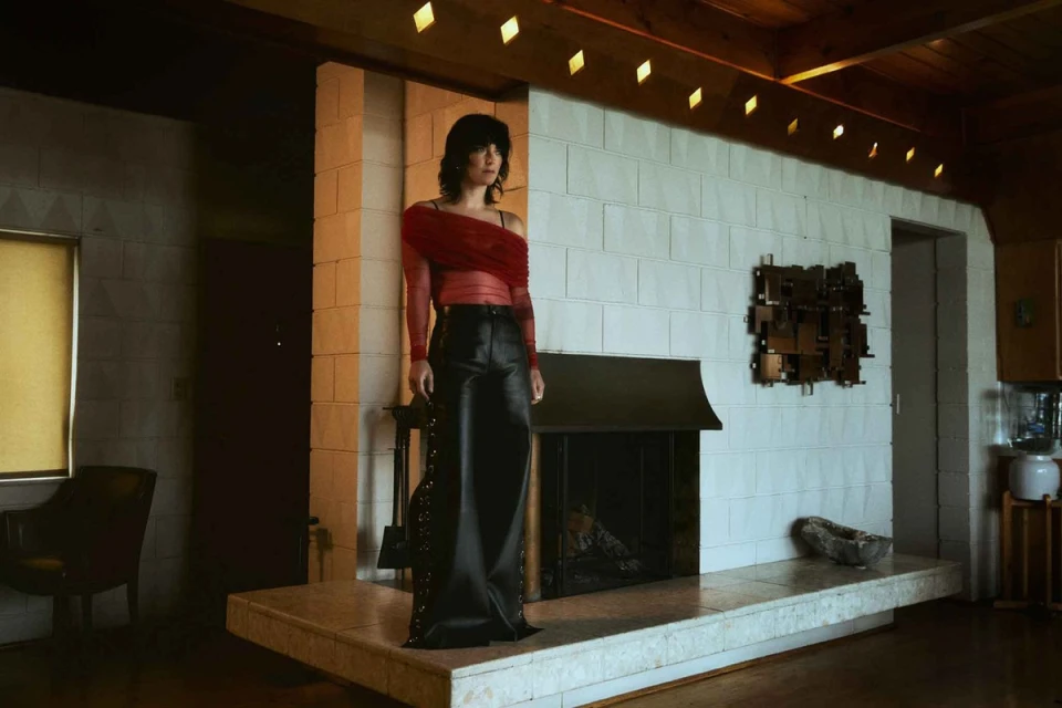 Adult woman posing in a sheer red blouse and black leather pants indoors, standing on a marble platform in front of a fireplace.