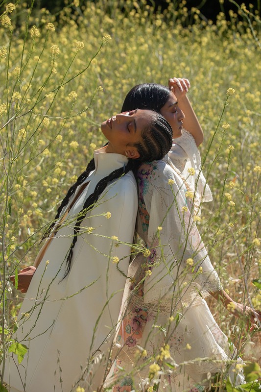 Two women stand back to back in a field of yellow wildflowers, wearing light-colored clothing, with an outdoor setting.
