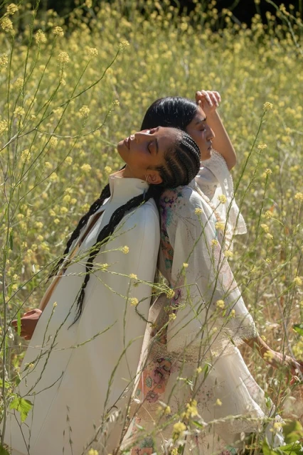 Two women stand back to back in a field of yellow wildflowers, wearing light-colored clothing, with an outdoor setting.